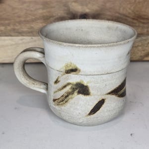 May include: A handmade ceramic mug with a cream-colored glaze and a curved handle. The mug features abstract brown and tan brushstrokes. The mug is a unique piece of pottery.