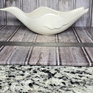 May include: A cream-colored ceramic bowl shaped like a stylized bird with a pointed beak and tail. The bowl features a smooth, glossy finish and a subtle embossed wing detail on the side. A metal Westcott ruler is placed in front, showing a length of approximately 12 inches.