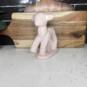 May include: A pink ceramic lamb figurine with a textured surface. The lamb is standing on a white surface.