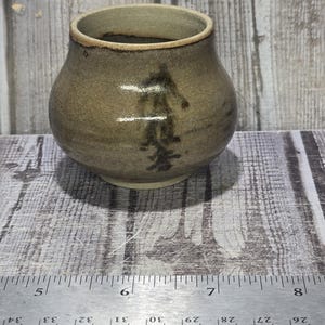 May include: A small, round, earth-toned ceramic pot with a wide opening and a dark, brush-stroke style design on the front. A metal ruler is positioned in front of the pot, showing it measures approximately 3 inches in width at its widest point.