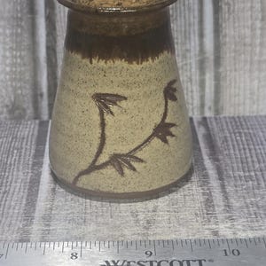 May include: A ceramic jar with a speckled beige body, a dark brown glazed rim, and a cork stopper. A dark brown branch design is painted on the front. A Westcott ruler placed in front shows the jar is approximately 3.5 inches in diameter.