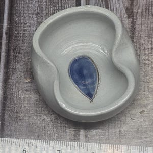 May include: A light gray ceramic bowl with an irregular, organic rim shape and a smooth, glossy finish. A single, dark blue, teardrop-shaped accent is centered at the bottom of the bowl. A metal ruler shows the bowl is approximately 3.5 inches wide.