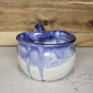 May include: A handmade ceramic bowl with a handle, featuring a white base and a blue, dripped glaze around the rim. The bowl has a rounded shape and a small handle. The background is a wooden surface.