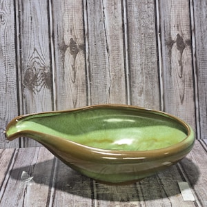 May include: A green and brown ceramic gravy boat with a spout. The interior is a lighter green, and the rim and exterior base are brown. The boat is oval-shaped and sits on a wooden surface.