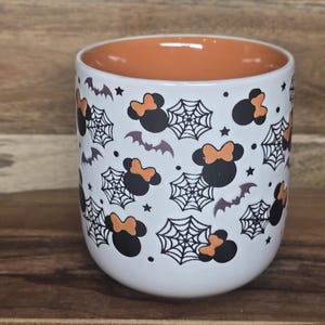 May include: White ceramic mug with an orange interior, decorated with black Minnie Mouse silhouettes with orange bows, spiderwebs, bats, and stars. The mug is a Halloween-themed item.