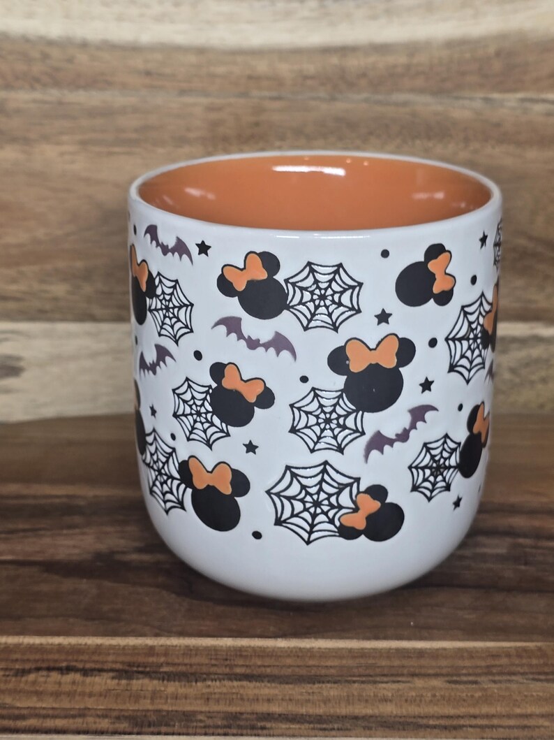 May include: White ceramic mug with an orange interior, decorated with black Minnie Mouse silhouettes with orange bows, spiderwebs, bats, and stars. The mug is a Halloween-themed item.