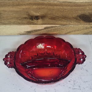 May include: A ruby red glass divided serving dish with ornate handles. The dish has a scalloped edge and a central divider. The Viking Hand Made USA logo is visible. The dish is on a white surface with a wooden background.
