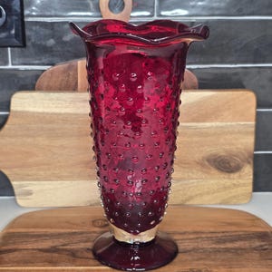 May include: A ruby red glass vase with a textured, dotted surface and a ruffled rim. The vase has a silver-colored base and is displayed on a wooden cutting board. The vase is a decorative item.