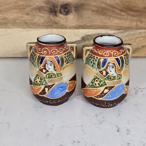May include: A pair of small, decorative Japanese vases. Each vase features a detailed painted design with a woman in traditional attire, set against a backdrop of gold and earth-toned colors. The vases have small handles and a brown base.