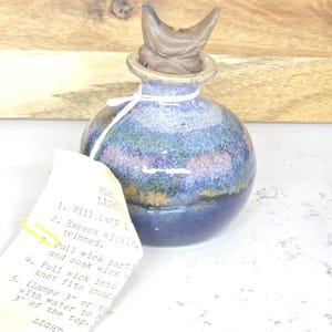 May include: A handcrafted, blue and purple ceramic oil lamp with a brown, decorative top. The lamp has a round body and a small opening. A tag with instructions is attached with a yellow clip. The lamp is on a white surface.