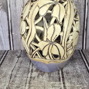 May include: A ceramic vase with a light cream-colored openwork floral pattern over a purple base. The vase features a narrow neck and a rounded body. A Westcott ruler placed at the base shows the vase stands approximately 9.5 inches tall.