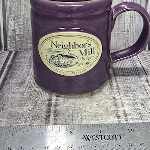 May include: A purple ceramic mug with a handle and a cream-colored oval decal featuring a mill illustration and the text Neighbor's Mill Bakery & Cafe Harrison, Arkansas. A metal Westcott ruler shows the mug is approximately 4 inches wide.