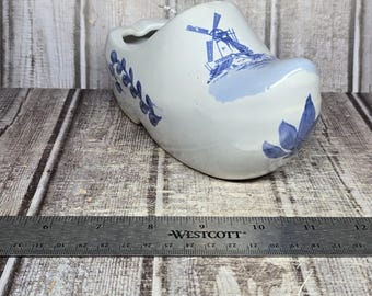 May include: A white ceramic clog with blue painted details, including a windmill illustration and floral patterns. A metal Westcott ruler is positioned in front, showing the object spans from approximately 6.5 inches to 10.5 inches in length.