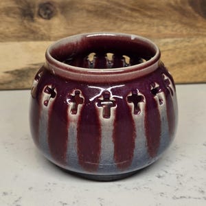 May include: A small, round ceramic pot with a deep burgundy glaze. The pot features cross-shaped cutouts around the sides and a lighter, almost white, glaze that creates vertical stripes. The rim is a lighter shade of burgundy.