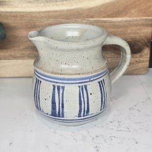 May include: A speckled ceramic pitcher with a handle and spout. The pitcher is off-white with blue vertical stripe detailing around the middle. The pitcher is sitting on a white surface.
