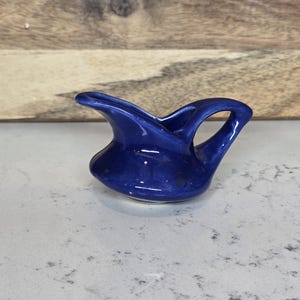 May include: A small, cobalt blue ceramic creamer with a handle and spout. The creamer has a glossy finish and sits on a white surface. The background is a wooden panel.