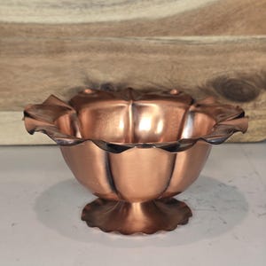 May include: A copper-colored bowl with a scalloped rim and a pedestal base. The bowl has a flower-like shape with curved petals. The metal has a shiny finish, reflecting light. The bowl is likely for decorative or serving purposes.