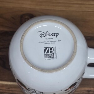 May include: White ceramic Disney mug with the Disney logo and text indicating it is dishwasher and microwave safe. The bottom of the mug also features the Zrike Brands logo and "Made in China."