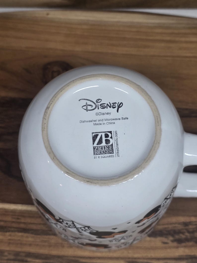 May include: White ceramic Disney mug with the Disney logo and text indicating it is dishwasher and microwave safe. The bottom of the mug also features the Zrike Brands logo and "Made in China."