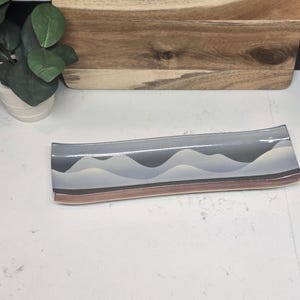 May include: A rectangular decorative glass tray with a wave design in shades of gray, white, and brown. The tray is placed on a white surface, with a wooden cutting board and a small potted plant in the background.