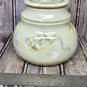 May include: A light cream-colored ceramic jar with a matching lid, featuring subtle blue and yellow painted accents and speckles. A metal ruler placed in front shows the jar has a diameter of approximately 6 inches at its widest point.
