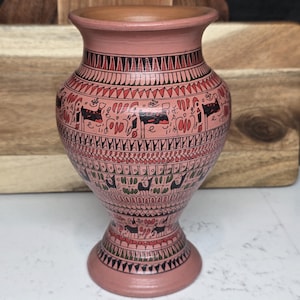 May include: A hand-painted, pink ceramic vase with a wide base and flared rim. The vase features intricate black and red tribal-style patterns and illustrations of animals and figures. The vase is approximately 6 inches tall.