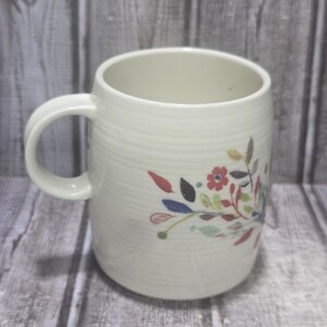 May include: Cream-colored ceramic mug with a floral design. The mug has a rounded shape with a handle and a textured, ribbed surface. The floral design features red, blue, and green leaves and flowers.