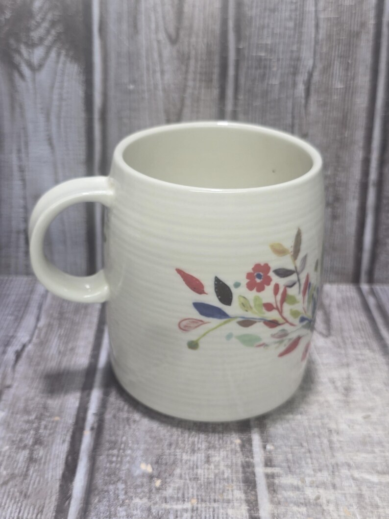 May include: Cream-colored ceramic mug with a floral design. The mug has a rounded shape with a handle and a textured, ribbed surface. The floral design features red, blue, and green leaves and flowers.