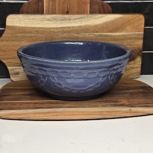 May include: A blue ceramic bowl with a decorative chain pattern around the rim. The bowl is set against a backdrop of wooden cutting boards, creating a rustic and functional aesthetic. The bowl is likely used for serving or food preparation.