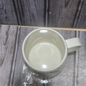 May include: A white ceramic coffee mug with a handle. The mug has a simple design and is empty. The mug is sitting on a wooden surface.
