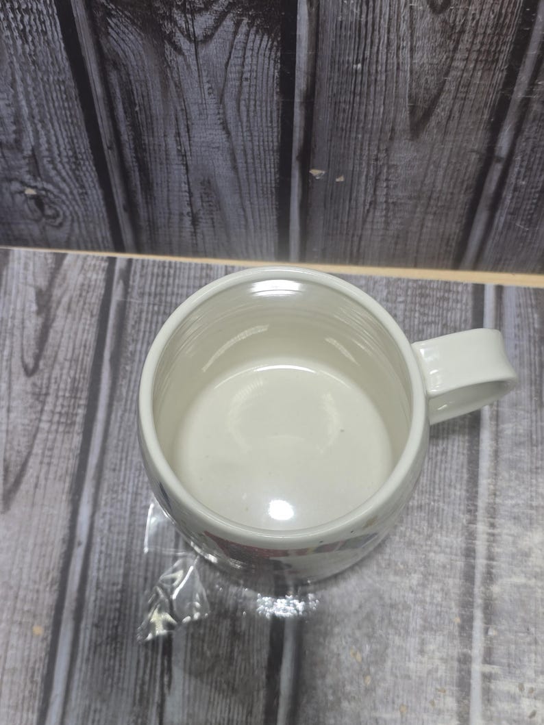 May include: A white ceramic coffee mug with a handle. The mug has a simple design and is empty. The mug is sitting on a wooden surface.