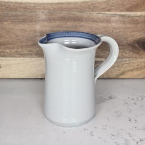 May include: White ceramic pitcher with a curved handle and a blue rim. The pitcher has a classic design and is suitable for serving beverages. The background is a wooden surface.