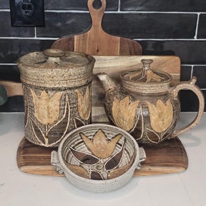 May include: A ceramic tea set with a teapot, lidded jar, and small bowl, all featuring a tulip design in shades of brown and tan. The set is displayed on a wooden cutting board, creating a rustic and earthy aesthetic. The background is a black tiled wall.