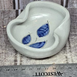 May include: A small, off-white ceramic bowl with an irregular, organic rim shape. The interior features three hand-painted blue leaves. A metal Westcott ruler is positioned in front, showing the bowl is approximately 3.5 inches in width.