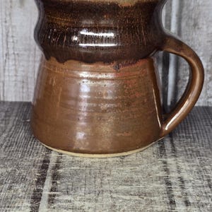 May include: A ceramic mug with a wide, flared rim and a tapered base. The mug features a glossy, dark brown glaze at the top that transitions into a lighter, earthy brown tone on the lower body. It includes a single curved handle on the right side.