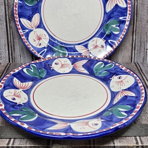 May include: Two round ceramic plates featuring a hand-painted blue background with white fish and green seaweed motifs. The rim is decorated with a red dotted pattern. A metal ruler placed underneath shows the plates are approximately 10 inches wide.