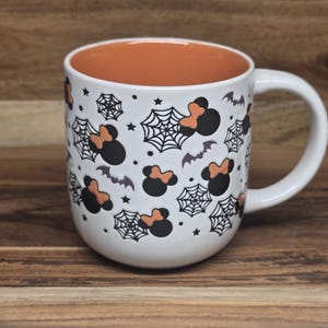May include: White ceramic mug with a Halloween theme. The mug features black and orange Minnie Mouse silhouettes, spiderwebs, bats, and stars. The interior of the mug is orange, and it has a white handle.