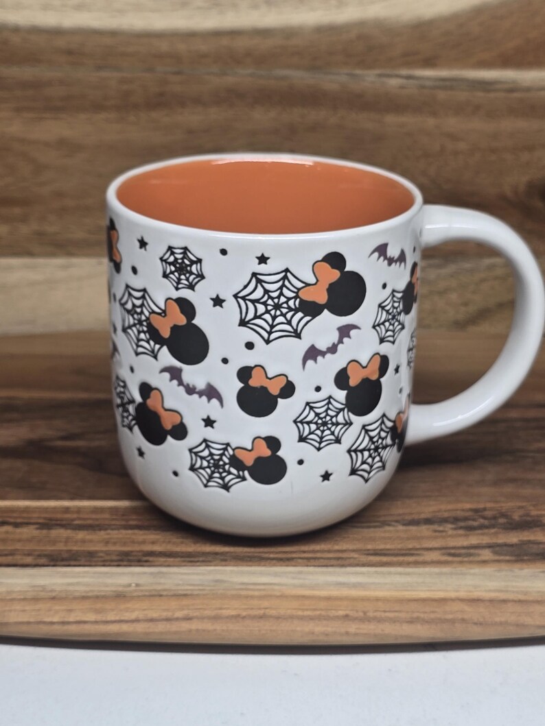 May include: White ceramic mug with a Halloween theme. The mug features black and orange Minnie Mouse silhouettes, spiderwebs, bats, and stars. The interior of the mug is orange, and it has a white handle.