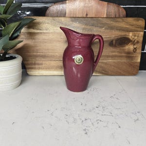 May include: A burgundy ceramic pitcher with a round, gold-colored sticker on the front. The pitcher has a handle and a spout.