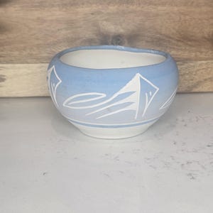 May include: A small, round ceramic bowl with a light blue exterior and white interior. The bowl features a white mountain design around the rim. The bowl is sitting on a white surface.