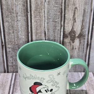 K&ouml;nnte beinhalten: Eine wei&szlig;e Keramiktasse mit gr&uuml;nem Innenraum und Henkel. Die Tasse hat ein festliches Design mit Mickey Mouse mit Weihnachtsm&uuml;tze und den Worten "Holiday Joy". Dekorative Schneeflocken sind ebenfalls vorhanden.