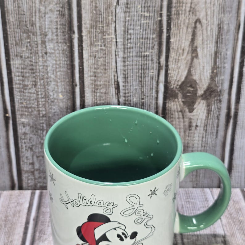 K&ouml;nnte beinhalten: Eine wei&szlig;e Keramiktasse mit gr&uuml;nem Innenraum und Henkel. Die Tasse hat ein festliches Design mit Mickey Mouse mit Weihnachtsm&uuml;tze und den Worten "Holiday Joy". Dekorative Schneeflocken sind ebenfalls vorhanden.