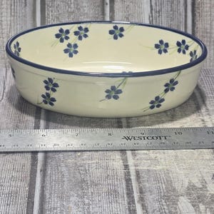 May include: A round, cream-colored ceramic bowl featuring a hand-painted pattern of small blue flowers with green stems. The bowl has a dark blue rim. A metal Westcott ruler is positioned in front, showing the bowl has a diameter of 6 inches.