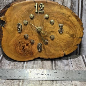 May include: A live edge wood slab wall clock featuring brass numerals 12, 3, 6, and 9, with brass dot markers for other hours and gold-tone hands. A Westcott ruler placed below shows the clock width is approximately 12 inches across.