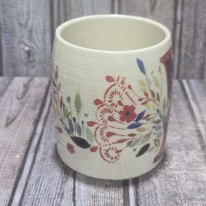 May include: Cream-colored ceramic mug with a floral design in red, blue, and green. The mug has a cylindrical shape and a wide opening. The floral pattern includes stylized flowers and leaves.