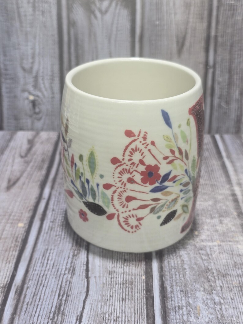 May include: Cream-colored ceramic mug with a floral design in red, blue, and green. The mug has a cylindrical shape and a wide opening. The floral pattern includes stylized flowers and leaves.