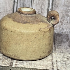 May include: A cylindrical tan ceramic oil lamp with a small side handle and a central opening at the top. A metal Westcott ruler is positioned in front of the object, showing the base diameter is approximately 3.5 inches wide.