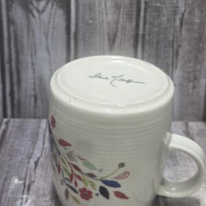 May include: White ceramic mug with a floral design in red, blue, green, and yellow. The mug has a curved handle and a textured surface. The bottom of the mug has the signature "Starke & Hoffman".
