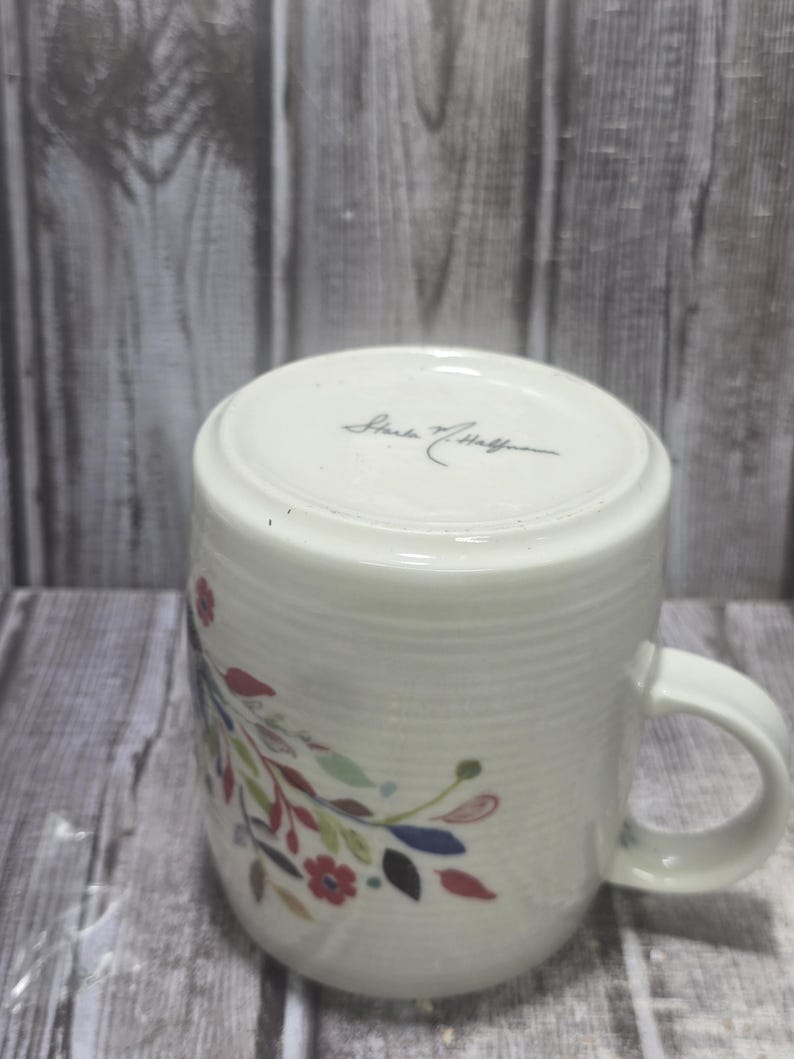 May include: White ceramic mug with a floral design in red, blue, green, and yellow. The mug has a curved handle and a textured surface. The bottom of the mug has the signature "Starke & Hoffman".
