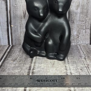 May include: A matte black ceramic figurine depicting two bears sitting together. The bears are positioned side-by-side with one arm around the other. A Westcott ruler is placed in front, showing the figurine is approximately 5.5 inches wide.